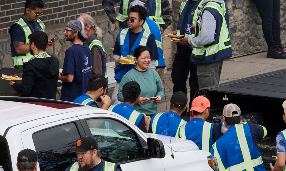 a large group of people are gathered in a parking lot for lunch. many of them wear blue and yellow safety vests