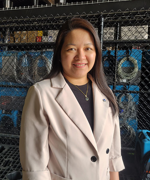 a woman wearing a black shirt with a white blazer and a thin gold chain with a "c" on a pendant stands in a warehouse for a photo