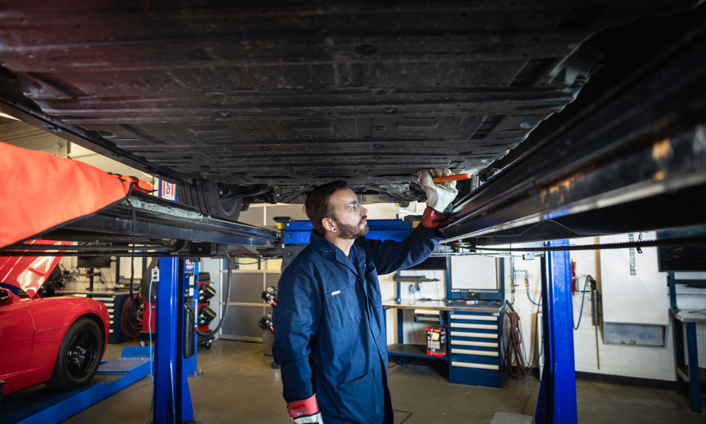 Tehničar u plavom kombinezonu radi ispod podignutog vozila na dizalu unutar auto radionice.
