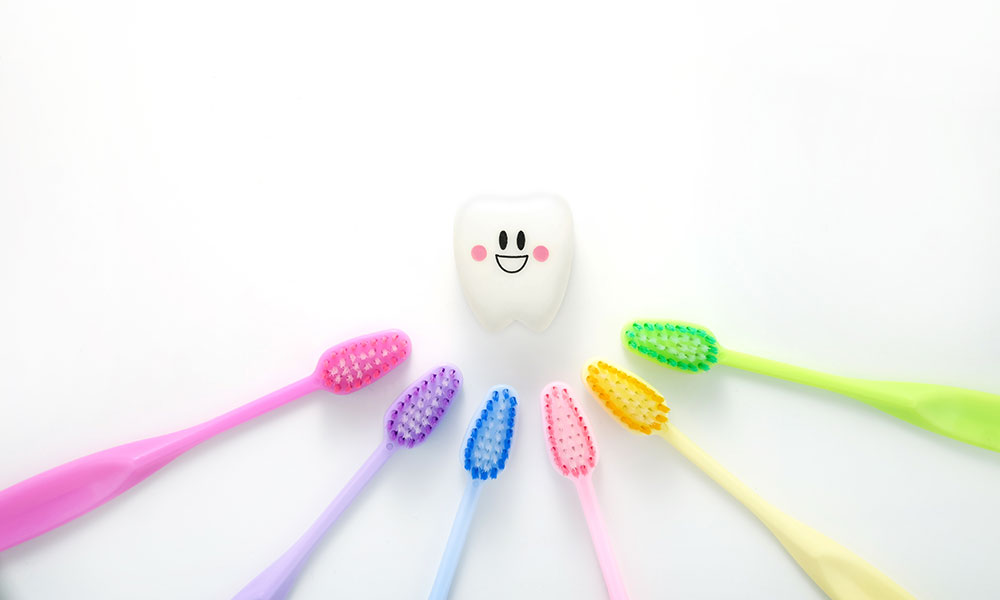 Flat lay of colorful toothbrushes arranged around a smiling tooth icon on a white background.