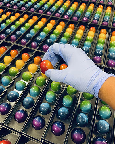 A gloved hand holds a red spherical object among rows of colorful, shiny, round chocolates in trays.
