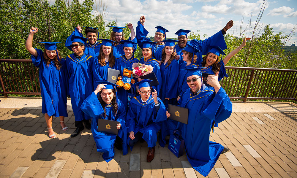 Group of people in blue graduation gowns and caps posing outdoors. Some hold diplomas and flowers, with several raising their arms in celebration. Trees and a partly cloudy sky are in the background.