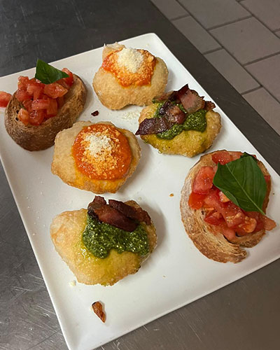A white rectangular plate with six pieces of bruschetta on a stainless steel surface. Three pieces have diced tomatoes and basil on sliced bread, two have red sauce and grated cheese on round bread bases, and one has green pesto, bacon, and grated cheese.