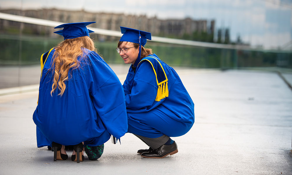 Two individuals in blue graduation gowns and caps squatting on a paved surface, with buildings and greenery blurred in the background.