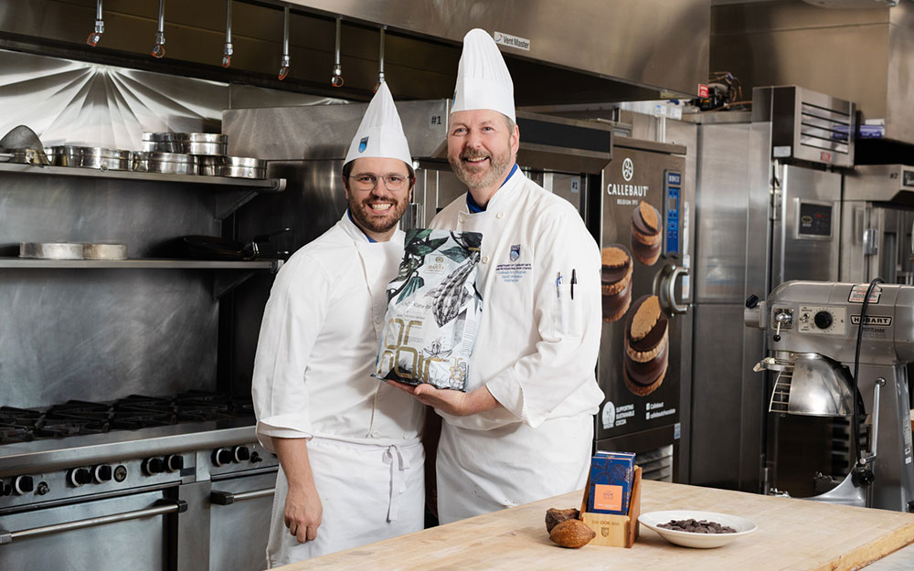 Two chefs in white uniforms smile and pose together in a professional kitchen while holding a bag of chocolate ingredients.