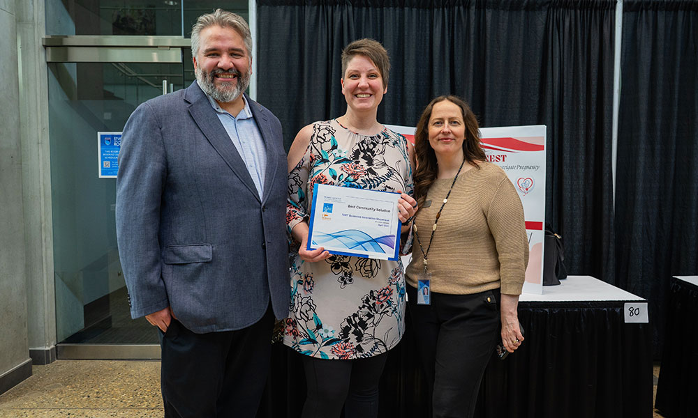 a photo of a man in a blue blazer, a woman in a white dress with a flower pattern, and a woman in a brown sweater. the woman in the dress, in the middle, holds a certificate.