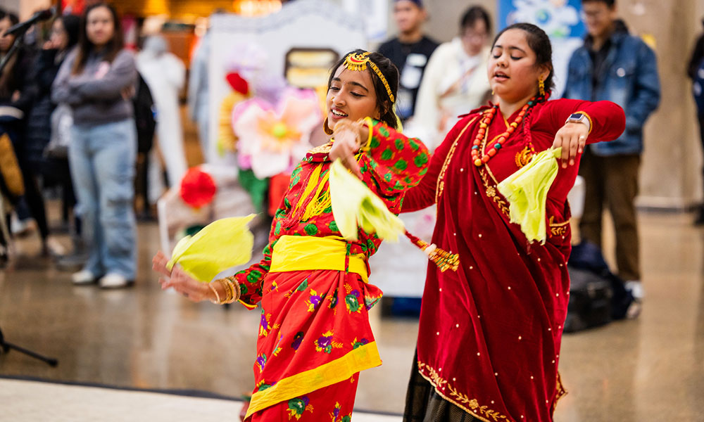 Two people wearing colorful traditional outfits performing a dance indoors with others in the background.