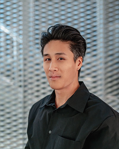 A person wearing a black collared shirt, standing in front of a metallic, patterned background.