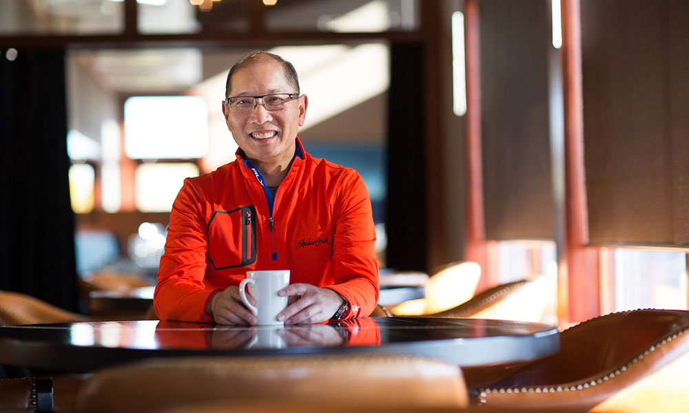 Person wearing a red jacket sits at a modern café table holding a white mug, with warm indoor lighting and blurred seating in the background.