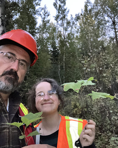 two people pose for a selfie in a forest. the man in the foreground wears a hard hat. they both are holding saplings.