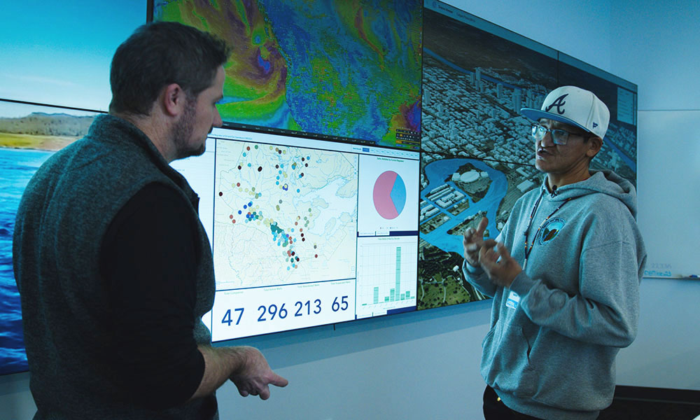Two people stand in front of a large wall of data screens, discussing maps, charts, and visual analytics.