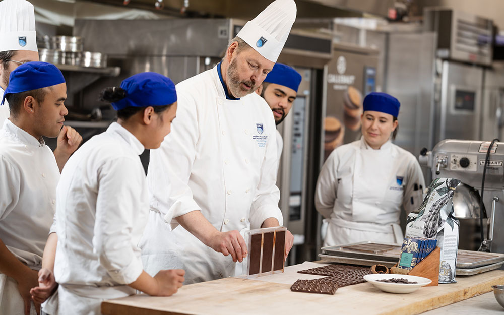 Chef instructor demonstrates chocolate bars to culinary students in a professional kitchen classroom.