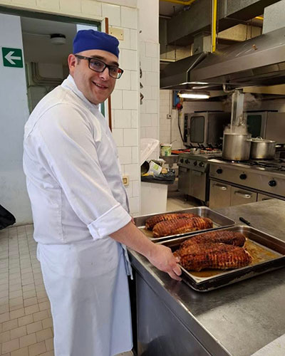 Person in a white chef's uniform and blue cap holding two trays of cooked meat in a professional kitchen with stainless steel appliances and an emergency exit sign on the wall.
