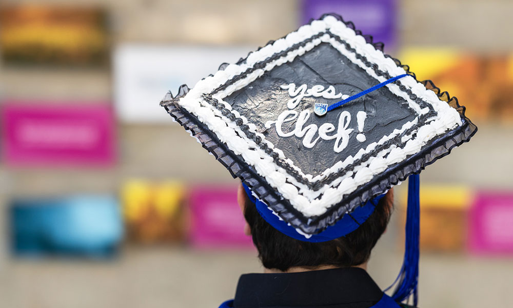 Back view of a person wearing a decorated graduation cap. The cap has white icing-like borders and the text 'yes, Chef!' on a black background. Colorful blurred signs or posters appear in the background.