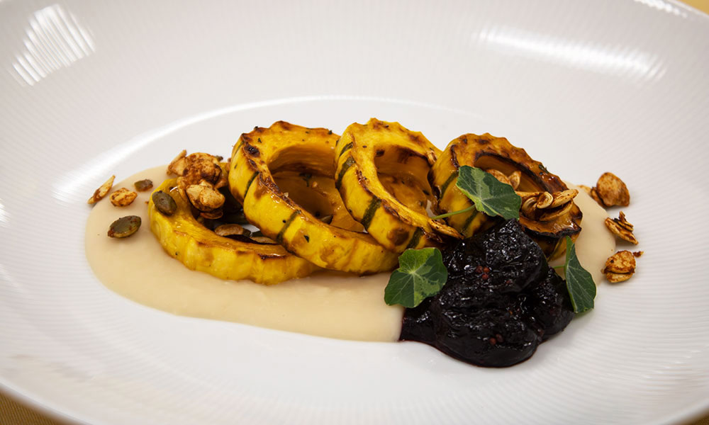 A plated dish features roasted delicata squash slices arranged with a light beige puree, a dollop of dark compote, granola, and small green leaves on a white plate.
