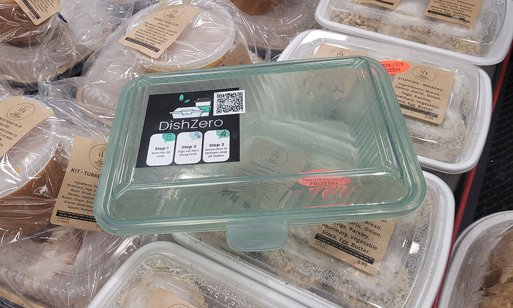 DishZero reusable plastic food container with green lid and QR code for zero-waste meal program, placed on packaged meal kits.
