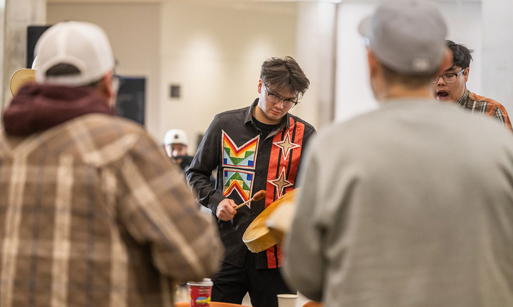 Drum group member playing a hand drum while people gather closely around during a round dance event.