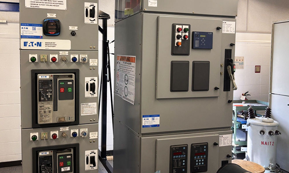 Large gray electrical control cabinets with switches, meters, and warning labels inside a technical training or industrial lab.