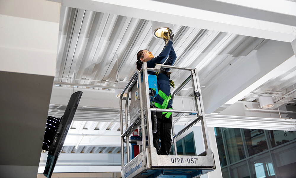 Person wearing safety gear on a scissor lift, working on a ceiling light fixture in an indoor space with exposed beams and multiple light fixtures.