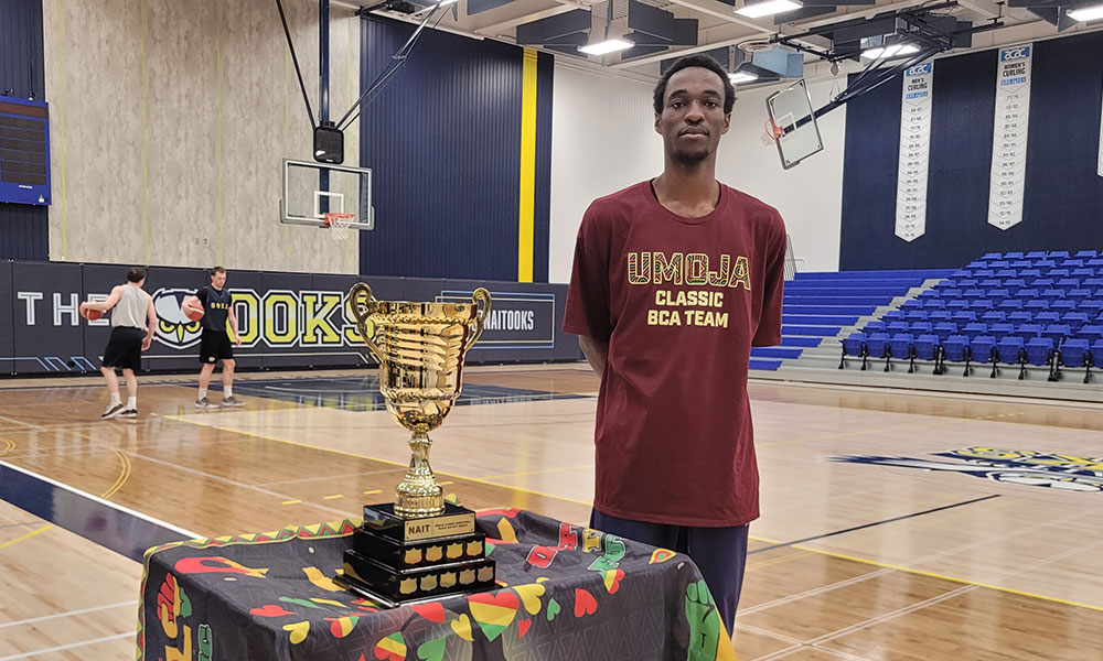 A basketball trophy sits on a colorful tablecloth in a gymnasium while a person in an “UMOJA Classic BCA Team” shirt stands nearby and players practice on the court in the background.