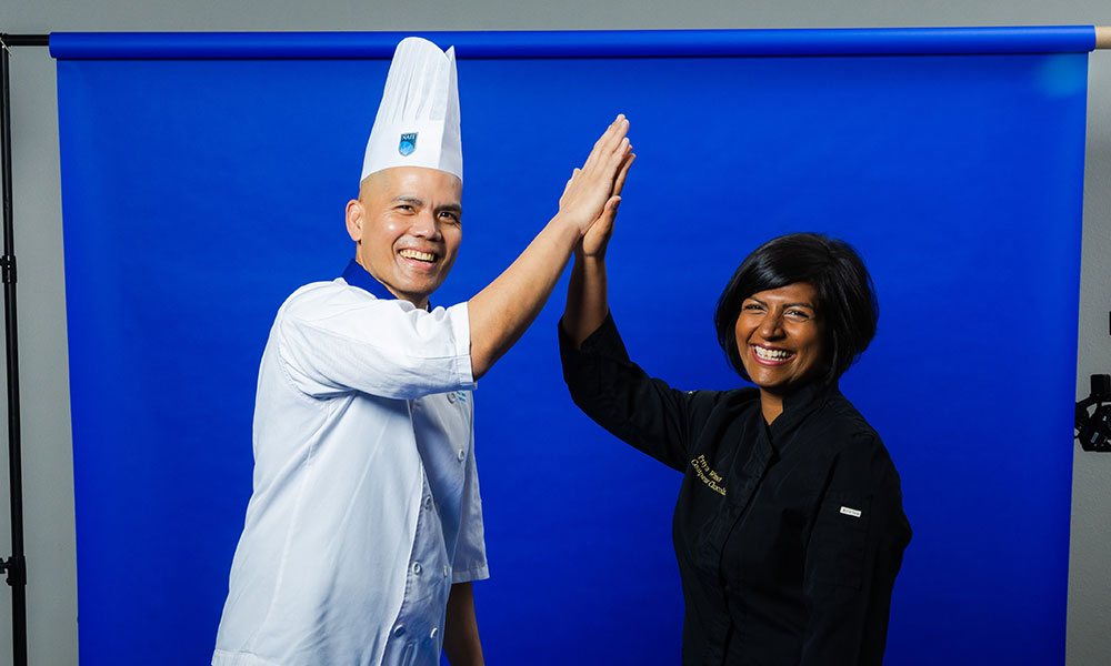 a man in chef's whites and a hat shares a high five with a woman in a black chef's jacket. they're in a photo studio in front of a blue background with its stand showing