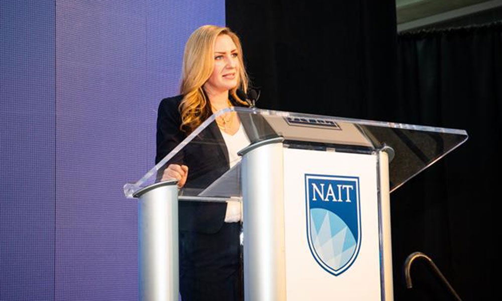 Person standing behind a clear podium with the NAIT logo, speaking at an event. A large screen and black curtain are visible in the background.