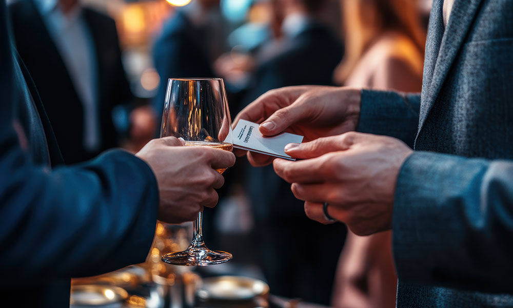 Two people exchanging business cards and holding champagne glasses at a crowded event.