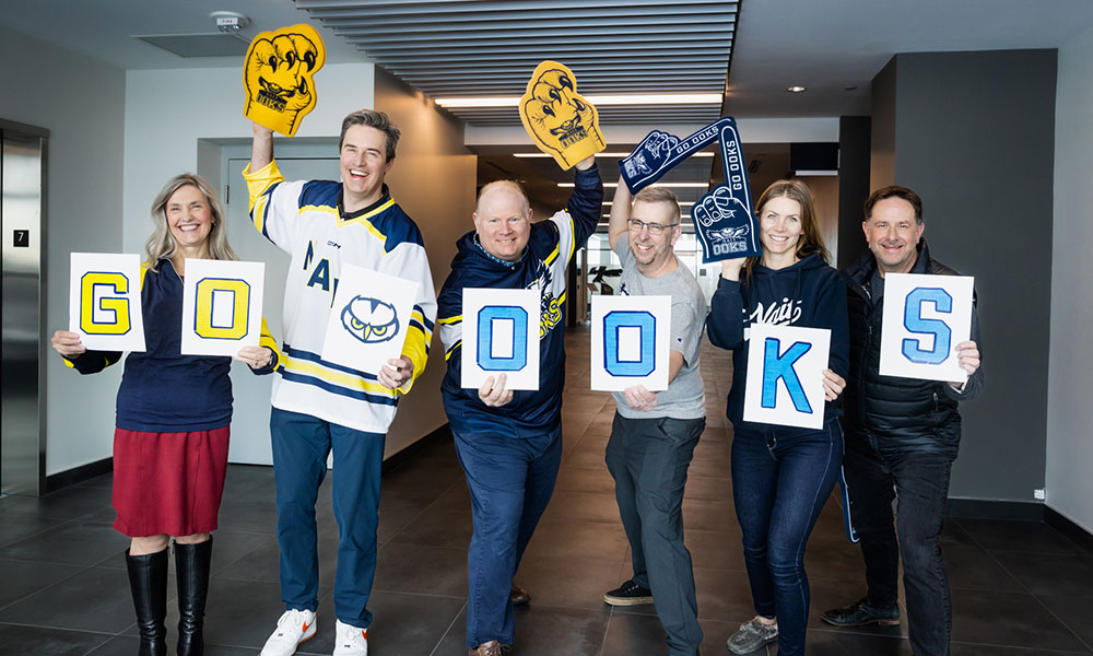 A group of people stand indoors holding letter signs and foam hands that spell out support for a sports team.
