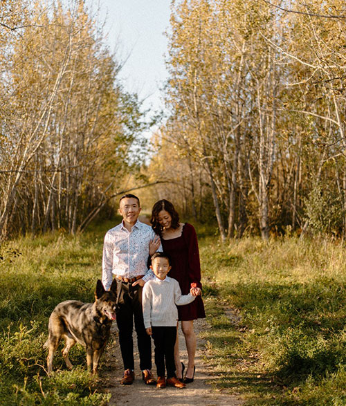 A serene scene of a family—two adults and one child—walking hand-in-hand along a wooded path, accompanied by a dog. The tall trees with sparse leaves suggest it's likely autumn.