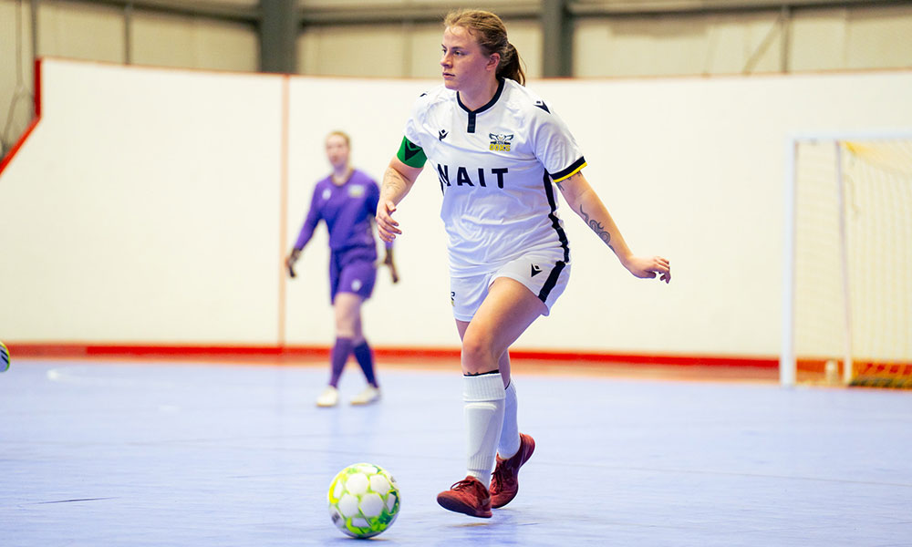 a woman in a white shirt with the word NAIT on the front moves along an indoor soccer pitch with the ball at her foot.