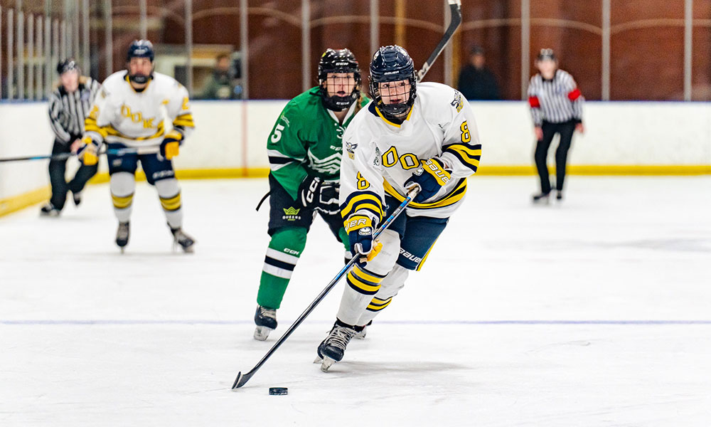 a female hockey player in a white uniform makes her way up ice with the puck. a player in a green uniform is chasing behind her.