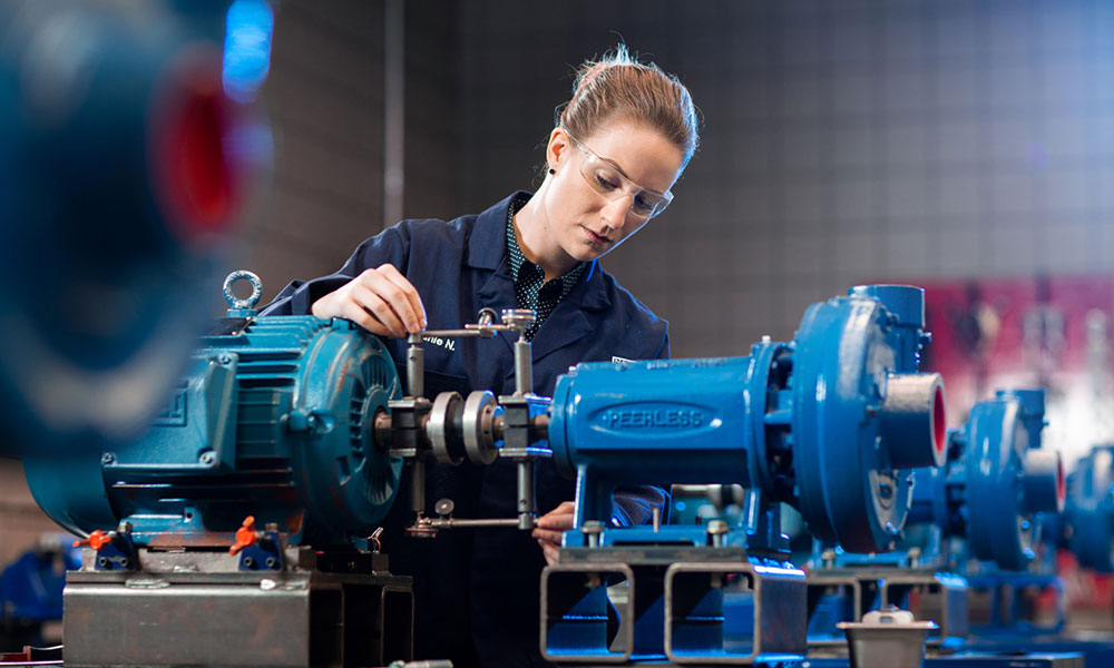 Person in a dark work uniform adjusting components on large blue industrial machinery, including motors and pumps, in a workshop setting.