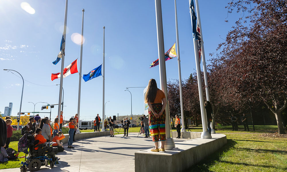 Group of people gathered around flagpoles with flags at half-mast on a sunny day, including individuals in orange shirts and one person in traditional Indigenous attire standing on a platform.