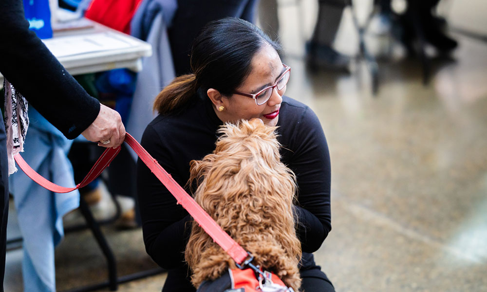 a woman with long dark hair and glass hugs a dog while someone holds it on a red leash