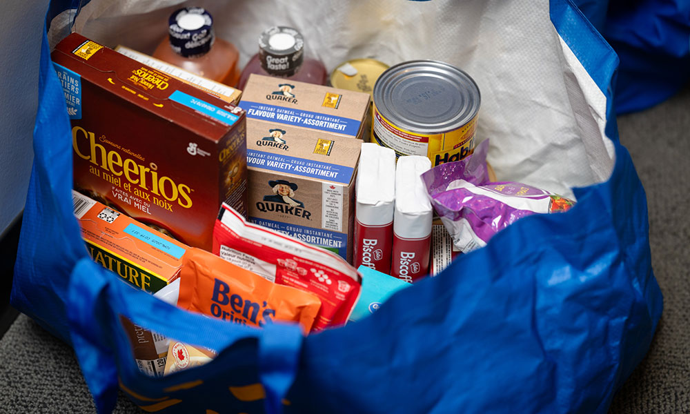 A blue reusable shopping bag filled with assorted packaged food items, including cereal, oatmeal, canned goods, rice, and snacks.
