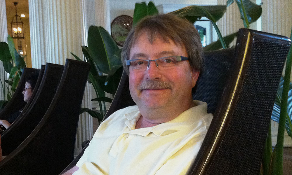 A person wearing glasses and a light-colored shirt sits in a chair with a relaxed posture in an indoor setting featuring large green plants and white columns. Another individual is seated in the background.