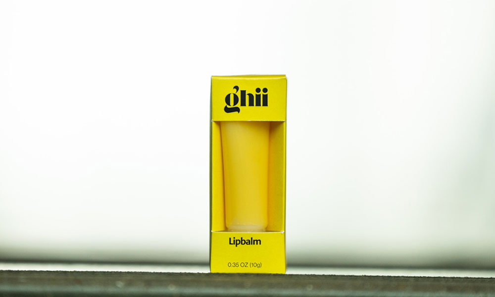 A single Ghii Lip Balm in a bright yellow tube is prominently displayed within its matching yellow cardboard box. The box has a rectangular cutout revealing the product, and features "ghii" in black text at the top, and "Lipbalm." The product is set against a plain white background.