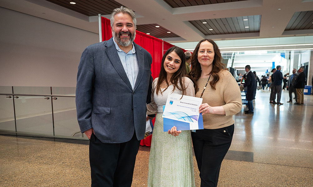 a photo of a man in a blue blazer, a woman in a white dress and a woman in a brown sweater. the woman in the dress, in the middle, holds a certificate.