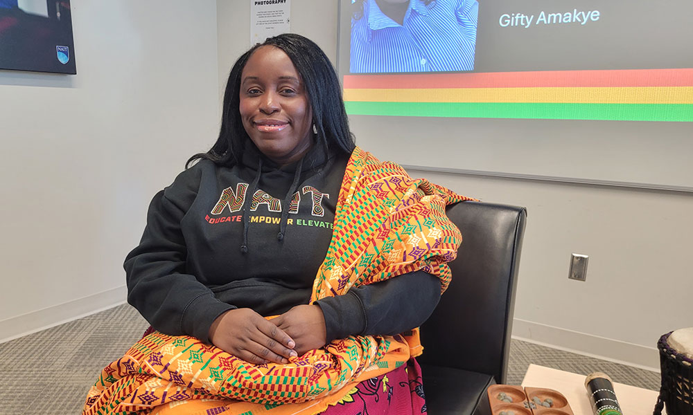 A person seated in a chair wearing a NAIT-branded hoodie and a vibrant patterned wrap, with a presentation screen in the background displaying the name “Gifty Amakye.”