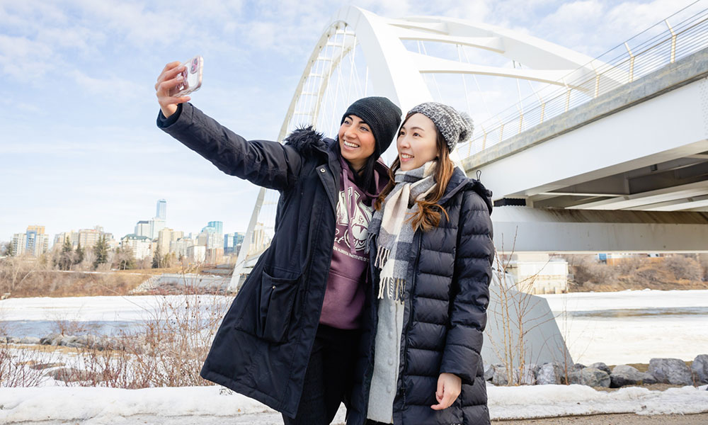 Two people in winter clothing taking a selfie near a bridge with a cityscape and partly cloudy sky in the background.