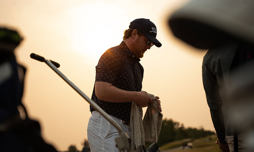 a male golfer holds cleans his clubs with a towel. he wears a dark t-shirt with white spots, a dark ball cap and sunglasses. the sun is rising behind him, giving the sky a soft yellow glow.