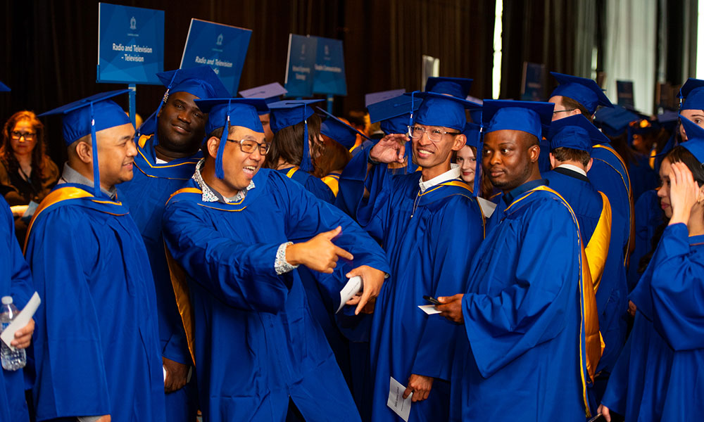 Group of graduates in blue caps and gowns gathered at a graduation ceremony. Some are holding papers or water bottles. Signs in the background read 'Radio and Television.' Faces are obscured.
