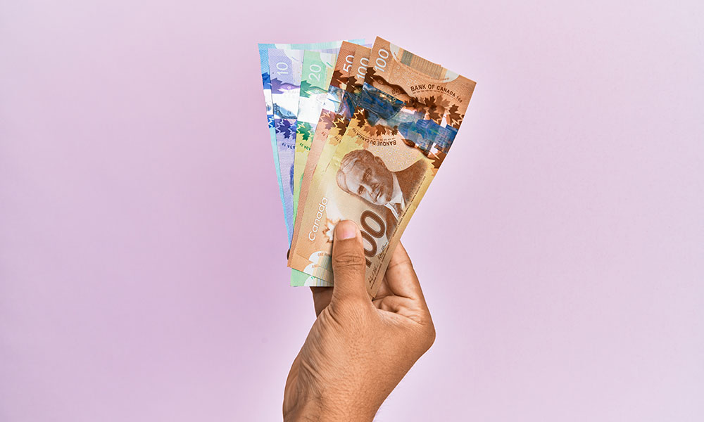 A hand holding a fan of Canadian banknotes against a light purple background.