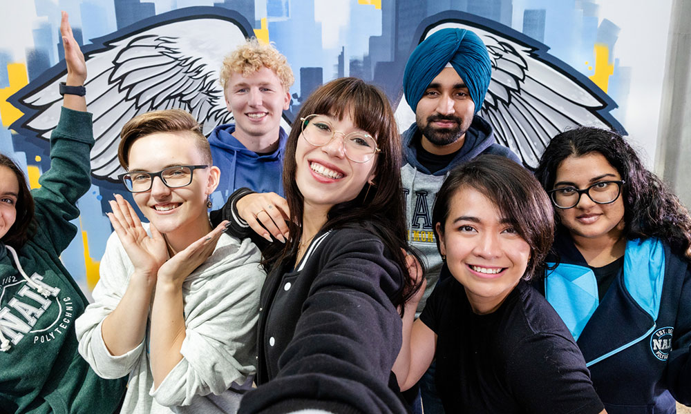 Group of six people posing for a selfie in front of a mural featuring angel wings.