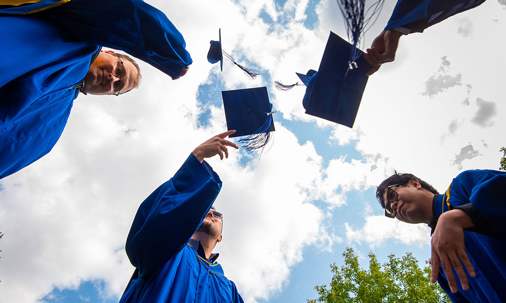 Group of people in blue graduation gowns and caps standing in a circle, throwing their caps into the air. The photo is taken from below, showing the sky with clouds.
