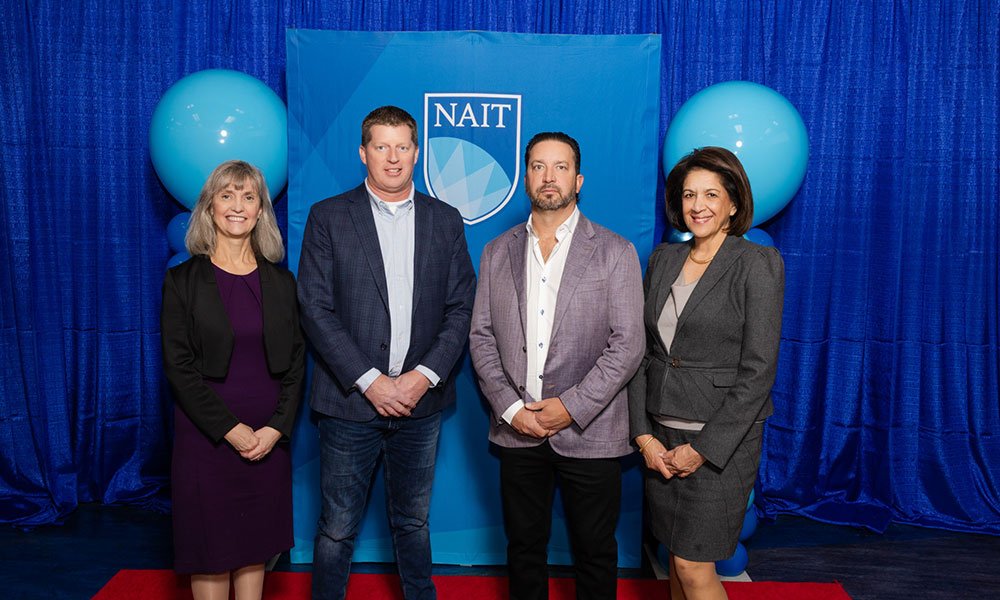 two women and two men between them pose for a photo. they are wearing business and formal attire. behind them is a blue background and blue balloons.