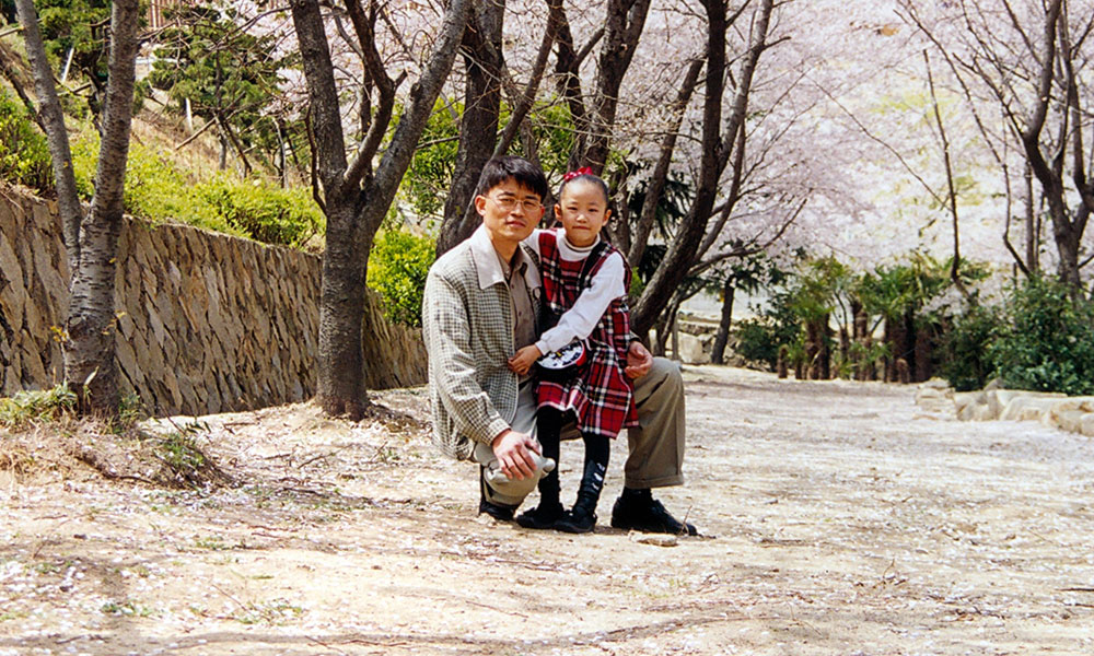 An adult kneeling beside a standing child in an outdoor setting with blossoming trees and a stone wall in the background. Both faces are blurred for privacy.