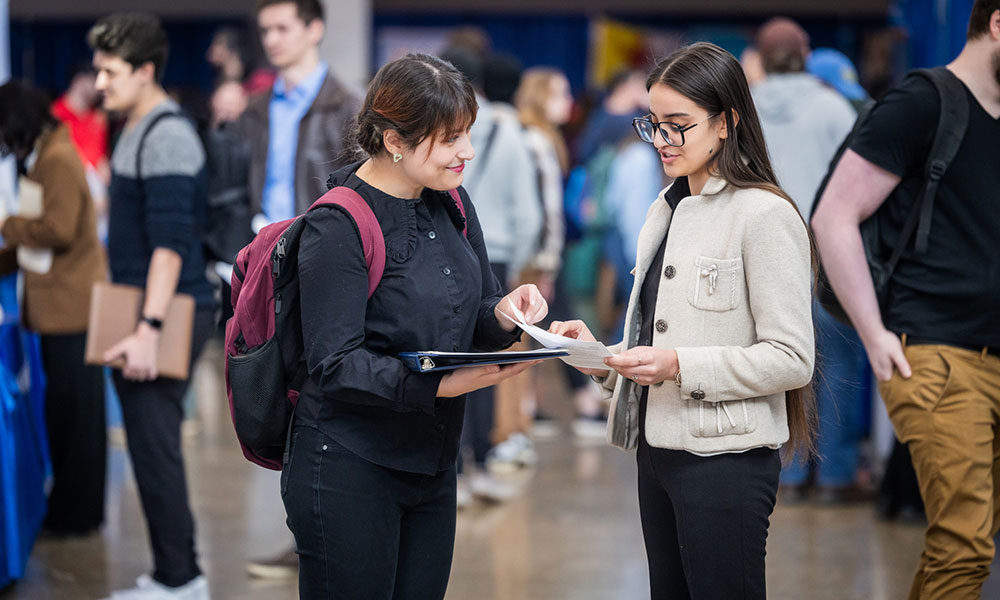 Two students review documents together at a busy hiring‑fair.
