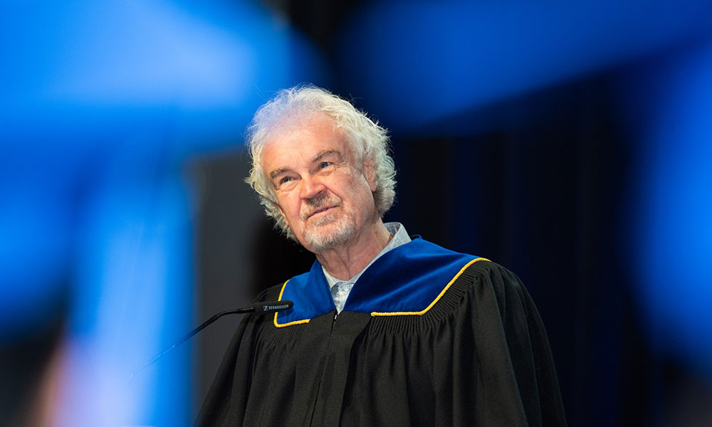 Person in academic regalia with a black gown trimmed in blue and gold, standing in front of a microphone. Background features blue and black colors.