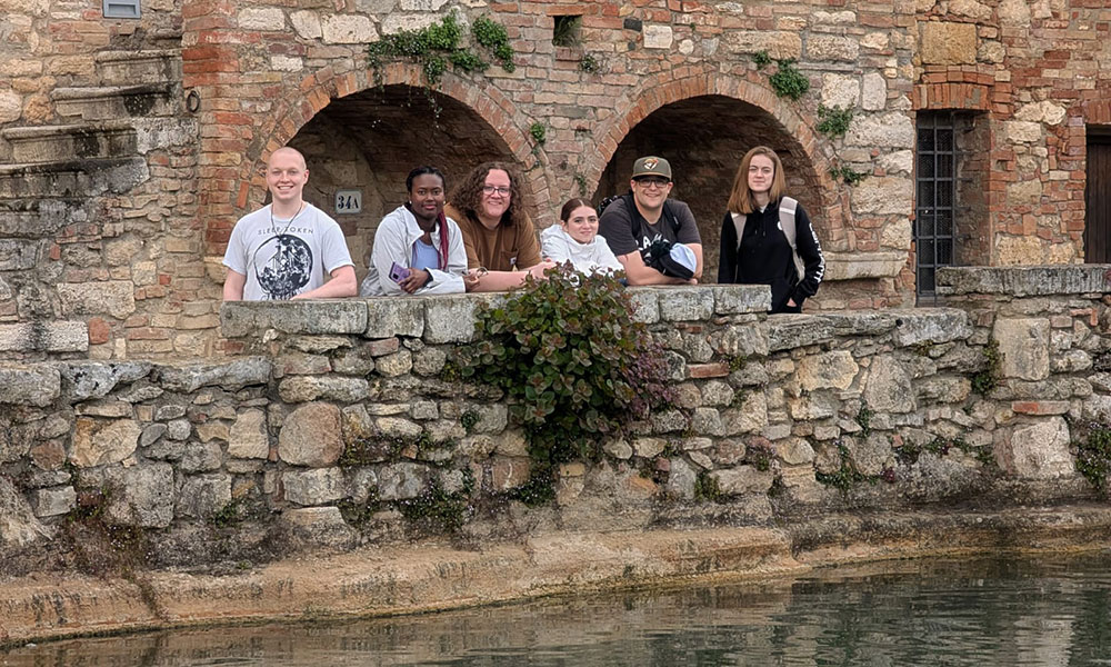 Group of six people standing behind a stone wall beside a body of water, with an old brick and stone building featuring arched openings and greenery in the background.